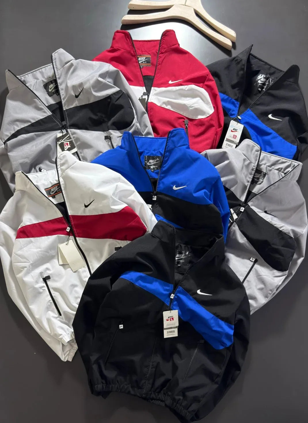 A group of Nike windbreaker jackets in multiple colorways, including red, black, white, and blue, arranged on a dark gray surface with wooden hangers visible at the top.