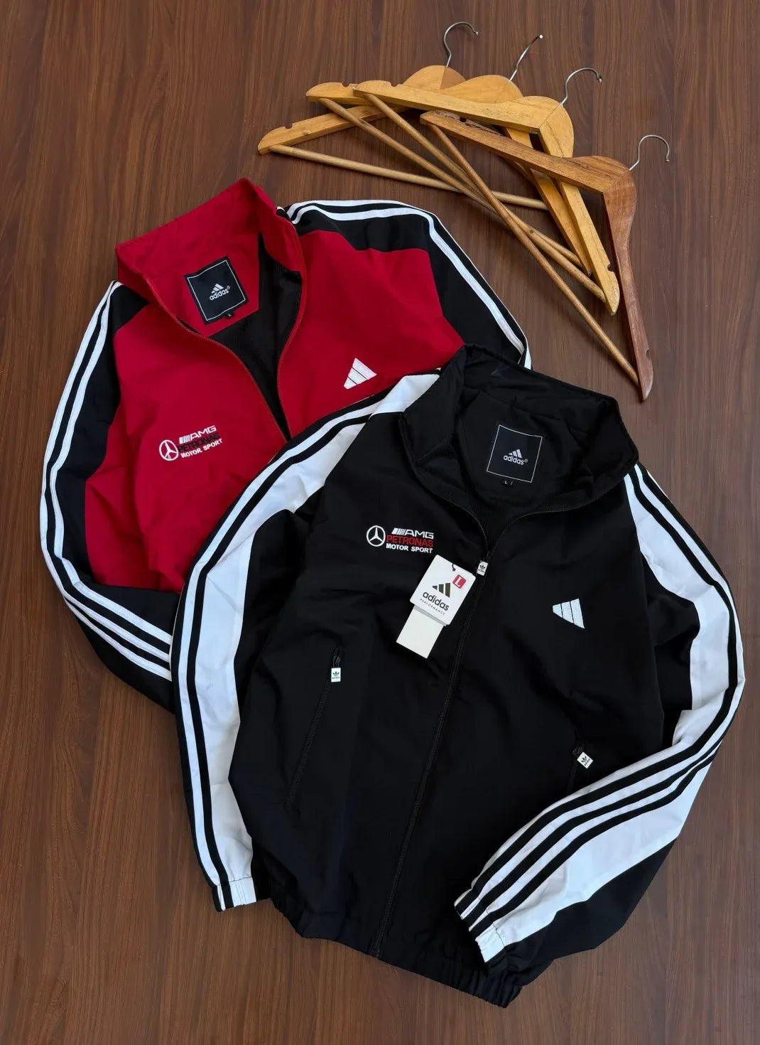 Two Adidas Mercedes-Benz track jackets, one black and one red, laid flat on a wooden surface with wooden hangers behind them. Both jackets feature the Mercedes-Benz logo and Adidas branding, with the black jacket in front displaying white three-stripe detailing on the sleeves.