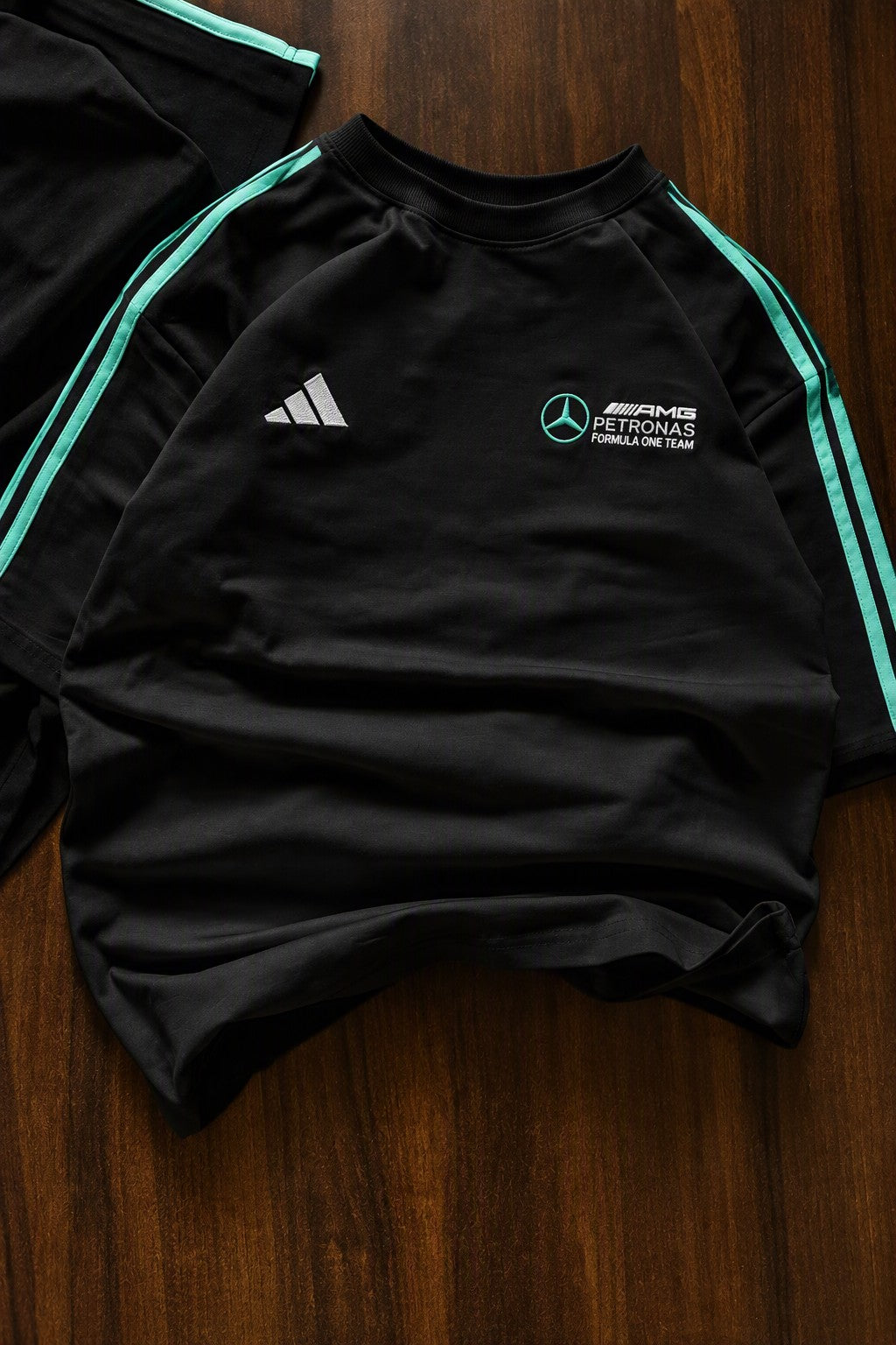 A black Adidas Mercedes-AMG Performance Racing long-sleeve top laid flat on a dark wood background, featuring mint green side stripes and white chest logos.