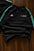 A black Adidas Mercedes-AMG Performance Racing long-sleeve top laid flat on a dark wood background, featuring mint green side stripes and white chest logos.