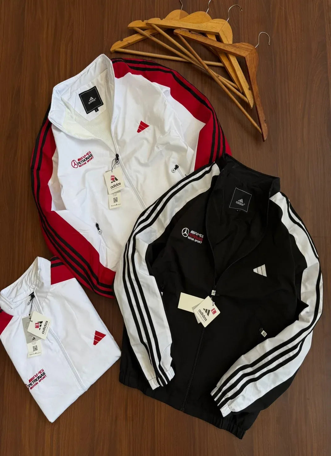Three Adidas track jackets laid flat on a wooden surface: one white with red side panels and black shoulder stripes, one black with white side panels and black shoulder stripes, and one folded white jacket. Wooden hangers are placed above the jackets.