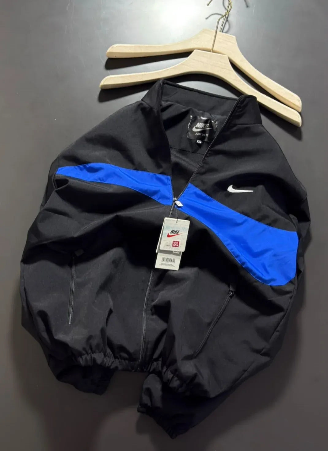 A black Nike windbreaker jacket with a bright blue diagonal stripe across the chest and a small white swoosh logo, hanging on a wooden hanger against a dark gray wall.