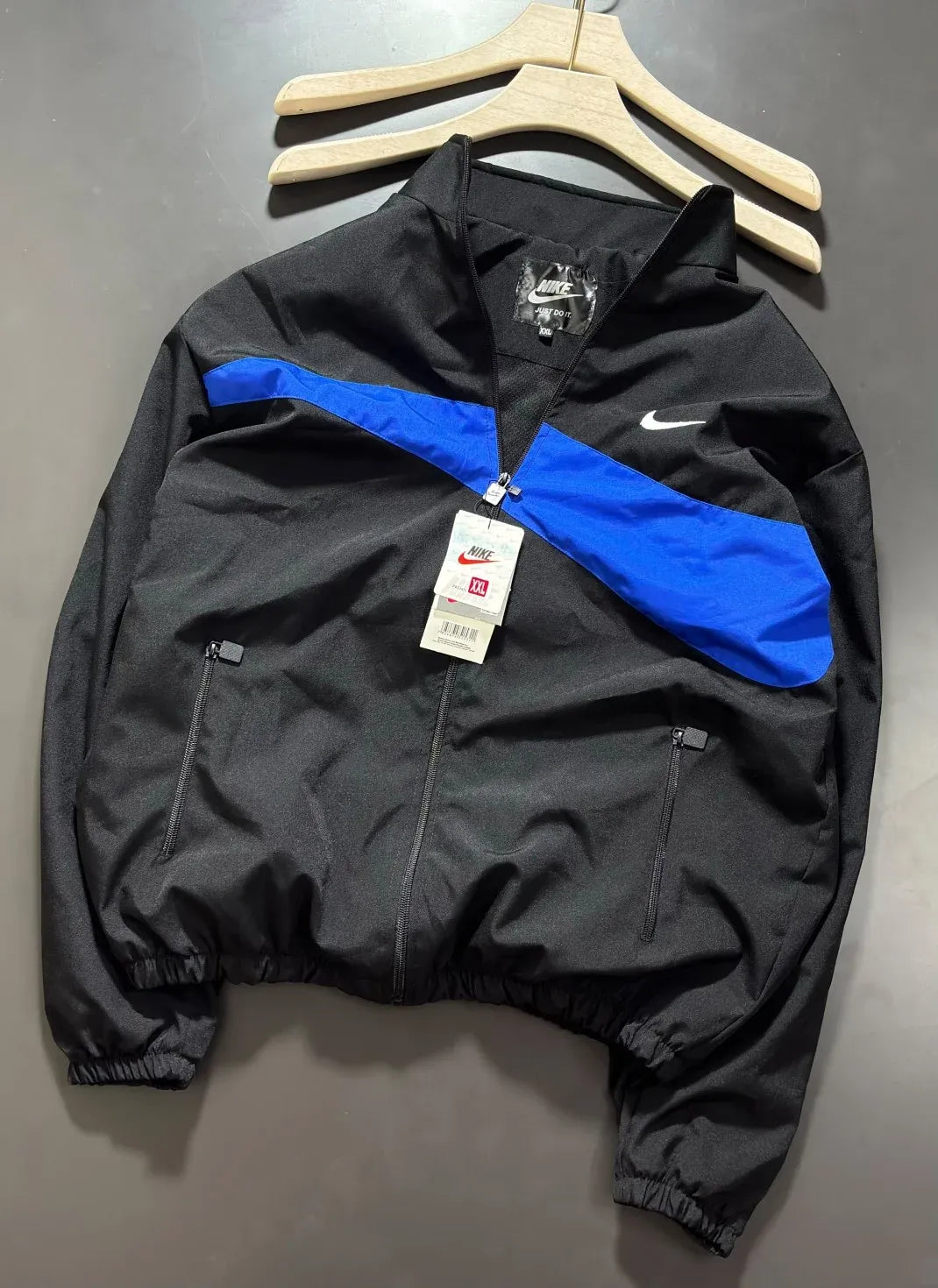A black Nike windbreaker jacket with a bold blue diagonal stripe across the chest, hanging on wooden hangers against a dark gray background. The jacket features a white Nike swoosh on the left chest, zippered pockets, and a hangtag still attached.