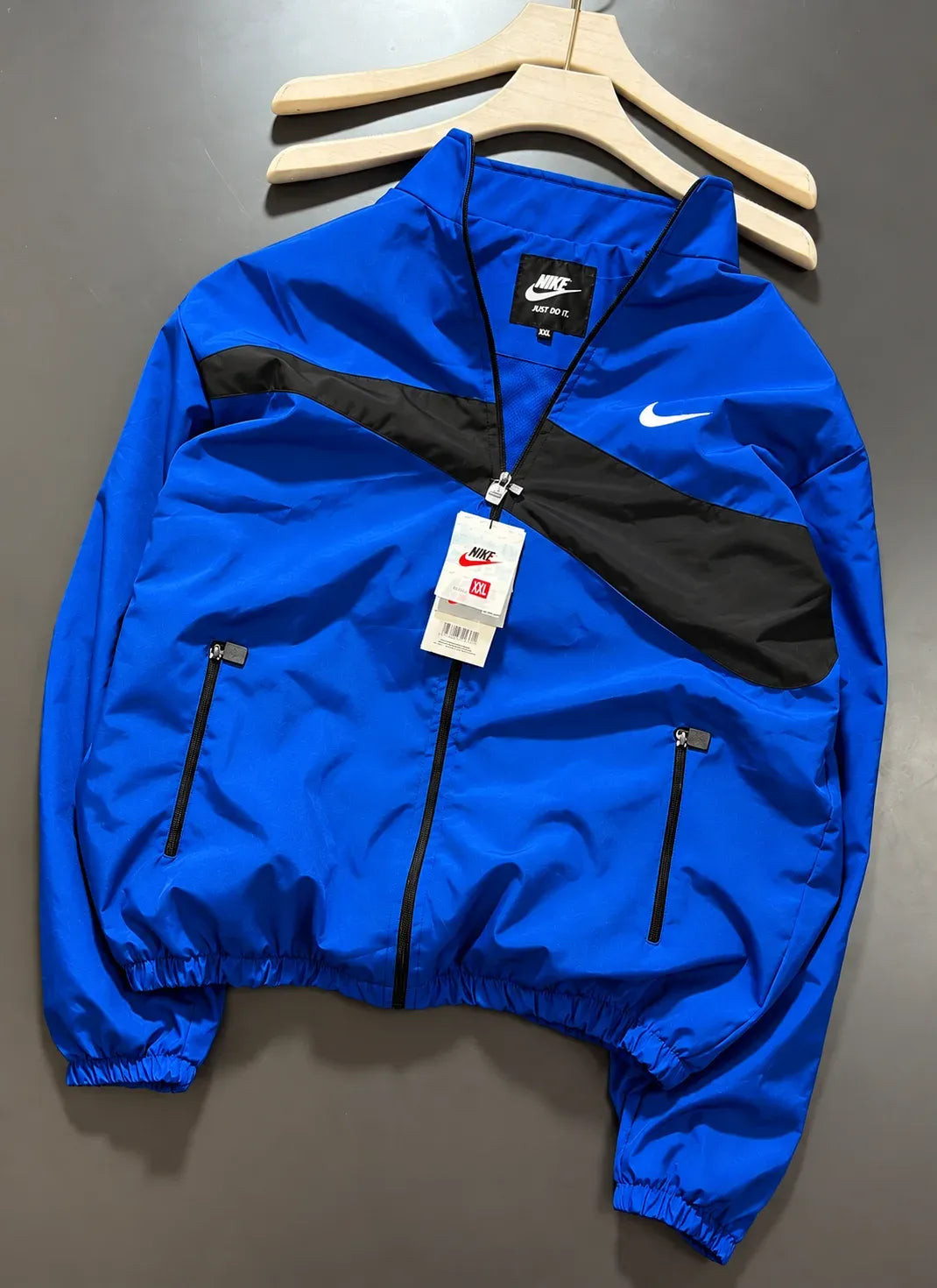A Nike windbreaker jacket in royal blue and black, hanging on a wooden hanger against a dark gray background. The jacket features a white Nike swoosh on the chest, a black Nike logo patch on the inner neck label, and a white hangtag attached to the zipper.