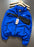 A Nike windbreaker jacket in royal blue and black, hanging on a wooden hanger against a dark gray background. The jacket features a white Nike swoosh on the chest, a black Nike logo patch on the inner neck label, and a white hangtag attached to the zipper.