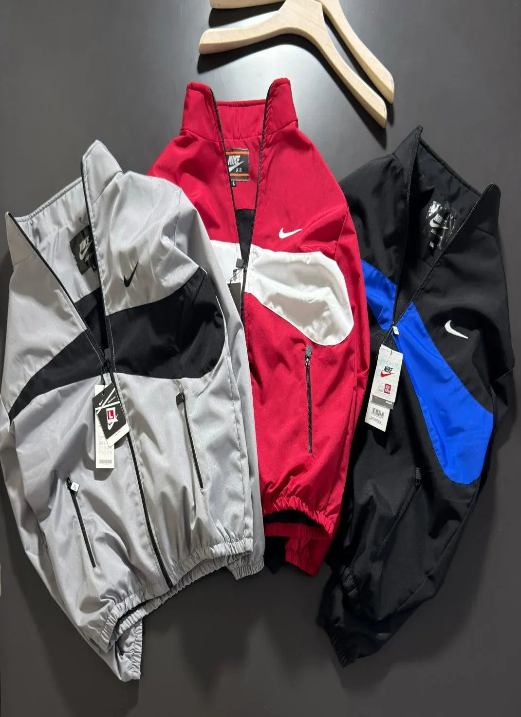 Three Nike windbreaker jackets laid flat on a dark gray surface: one silver with black panels, one red with white panels, and one black with blue panels. A wooden hanger is partially visible at the top.