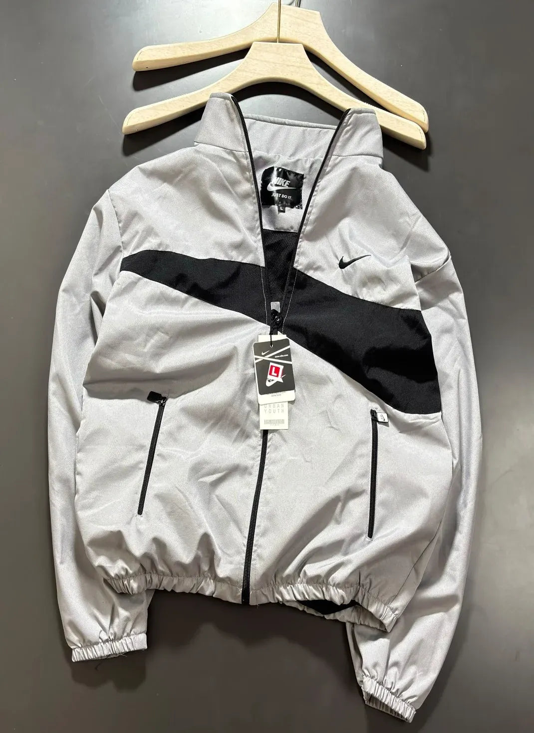 A light gray Nike windbreaker jacket with a black diagonal chest stripe and black zippers, hanging on a wooden hanger against a dark gray wall.