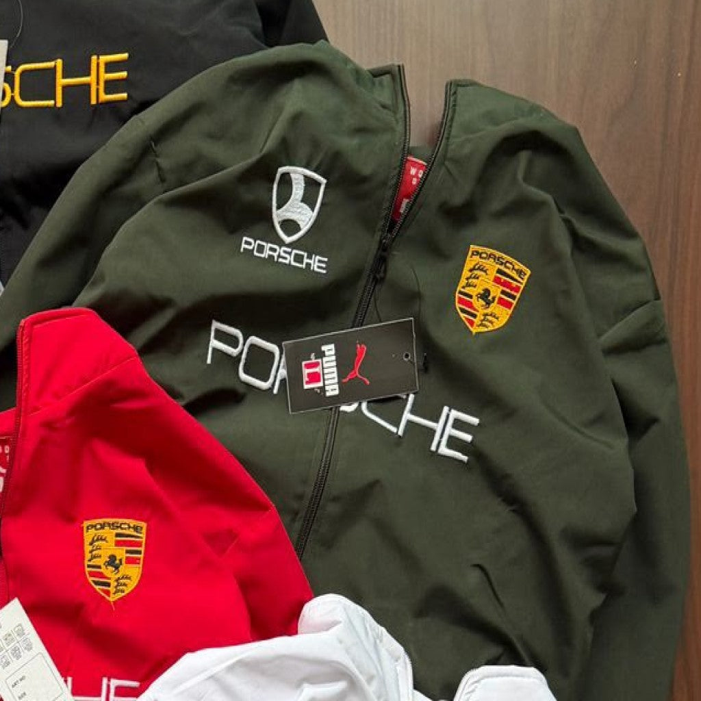 A dark olive green Porsche zip-up jacket laid flat on a wood surface, with a white Porsche crest logo on the chest, a yellow and red Porsche crest patch on the sleeve, and a black hangtag attached. Partially visible behind it are red and black Porsche jackets.