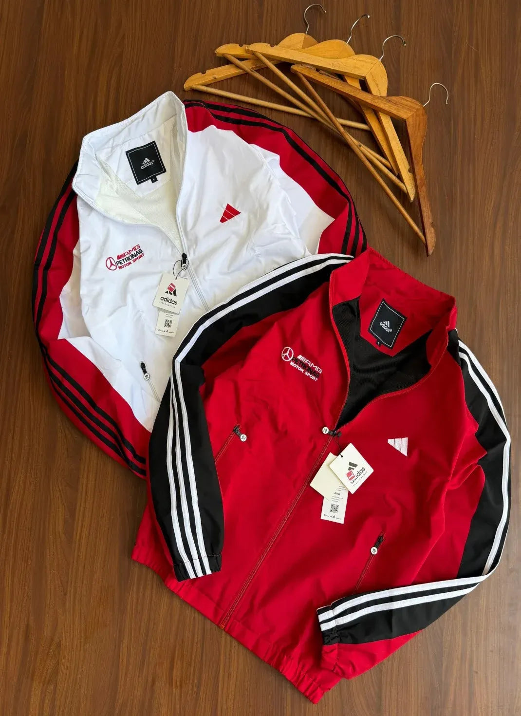 Two Adidas track jackets laid flat on a wooden surface, one white with red and black stripes and one red with black and white stripes, with wooden hangers above them.