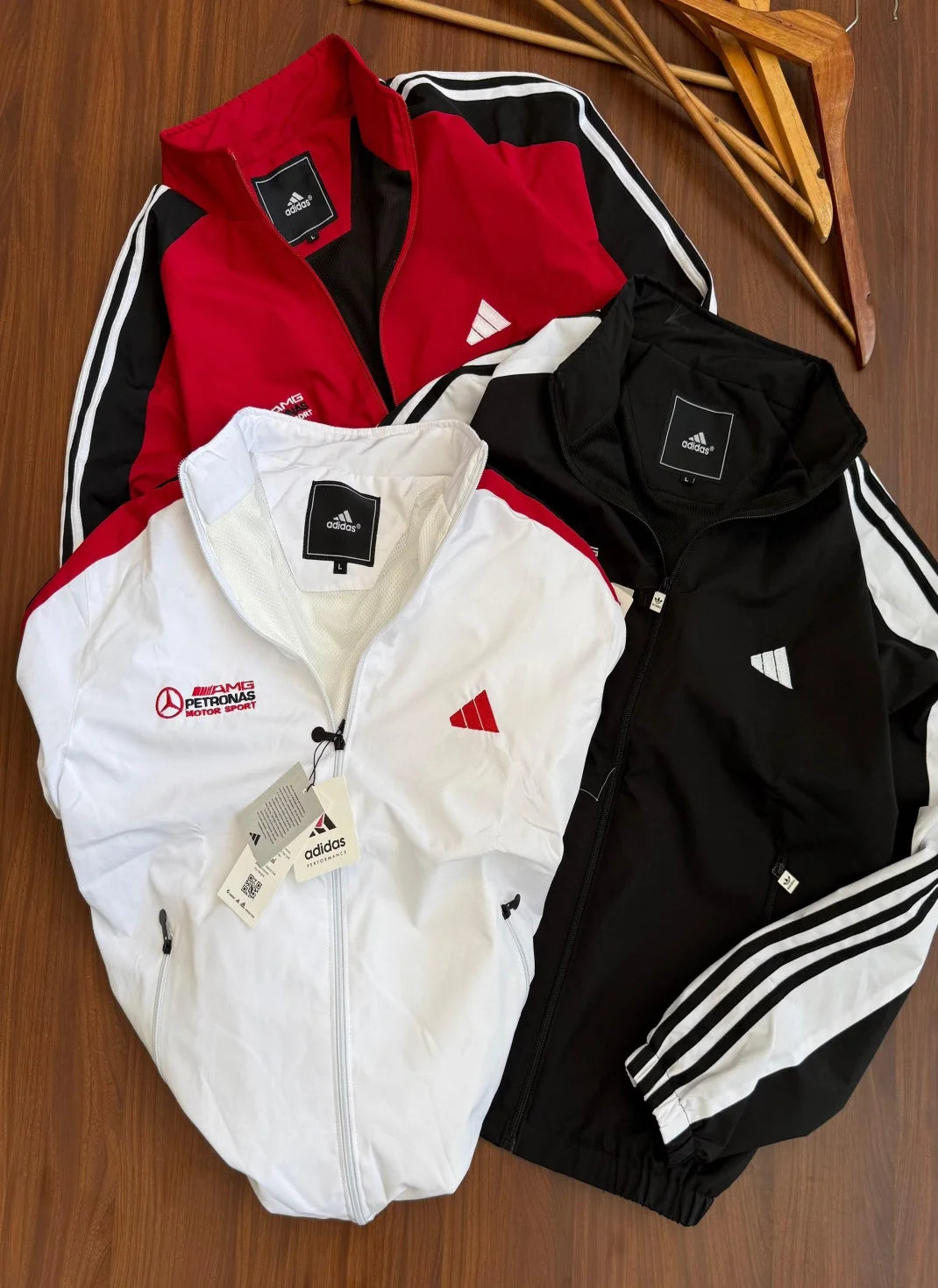 Three Adidas track jackets in red, white, and black are laid out on a wooden surface. The jackets feature the Adidas brand logo, a Mercedes-Benz Top Gear patch, and three stripes down the sleeves.