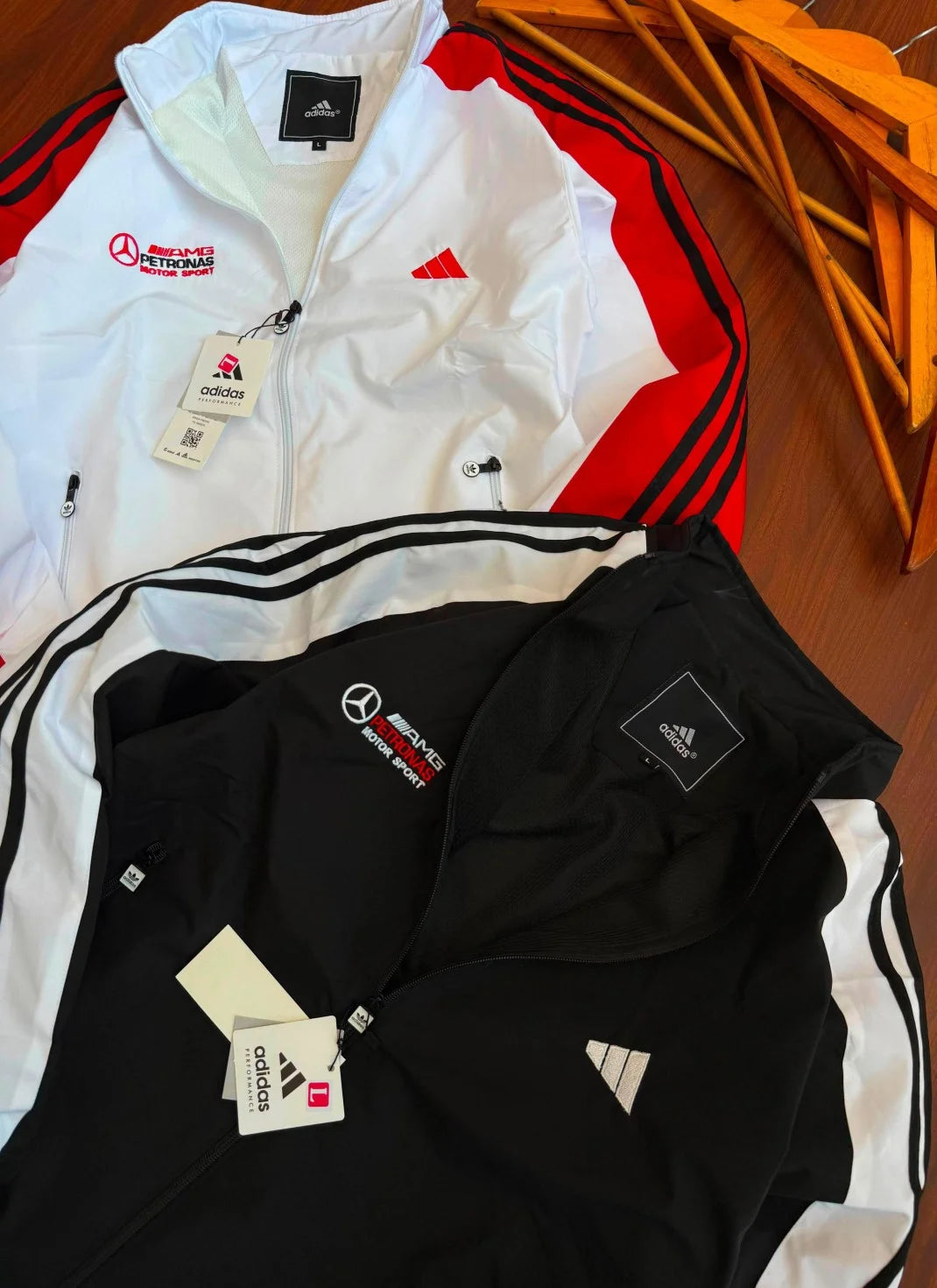 Two Adidas track jackets laid flat on a wooden surface: one white with red side panels and black stripes, and one black with white side panels and black stripes. Both jackets show Mercedes-Benz and Adidas branding, and hangtag tags are attached.