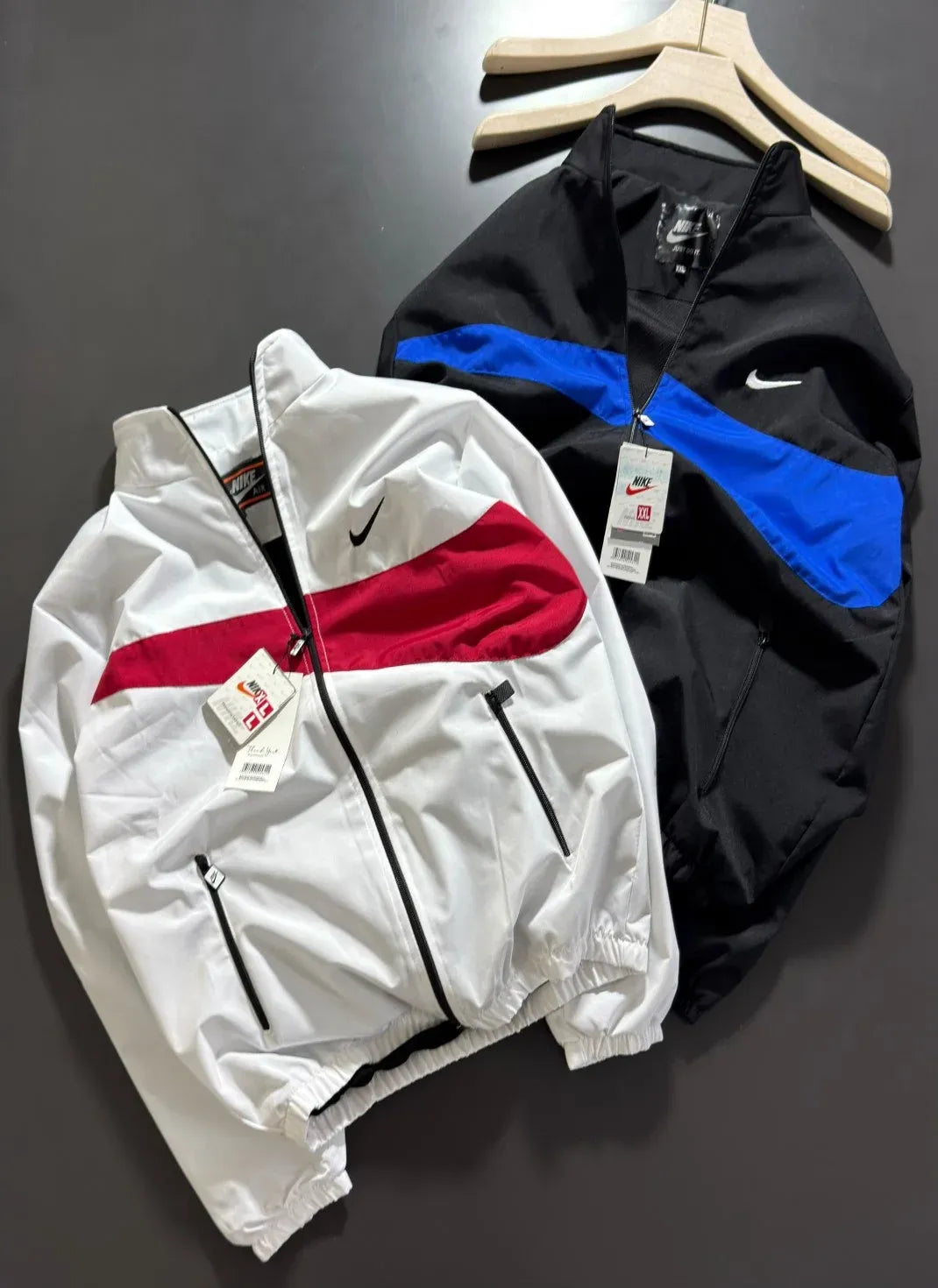 Two Nike windbreaker jackets laid flat on a dark gray surface, one white with a red chest stripe and one black with a blue chest stripe. Both jackets feature a white Nike swoosh and hang tags.