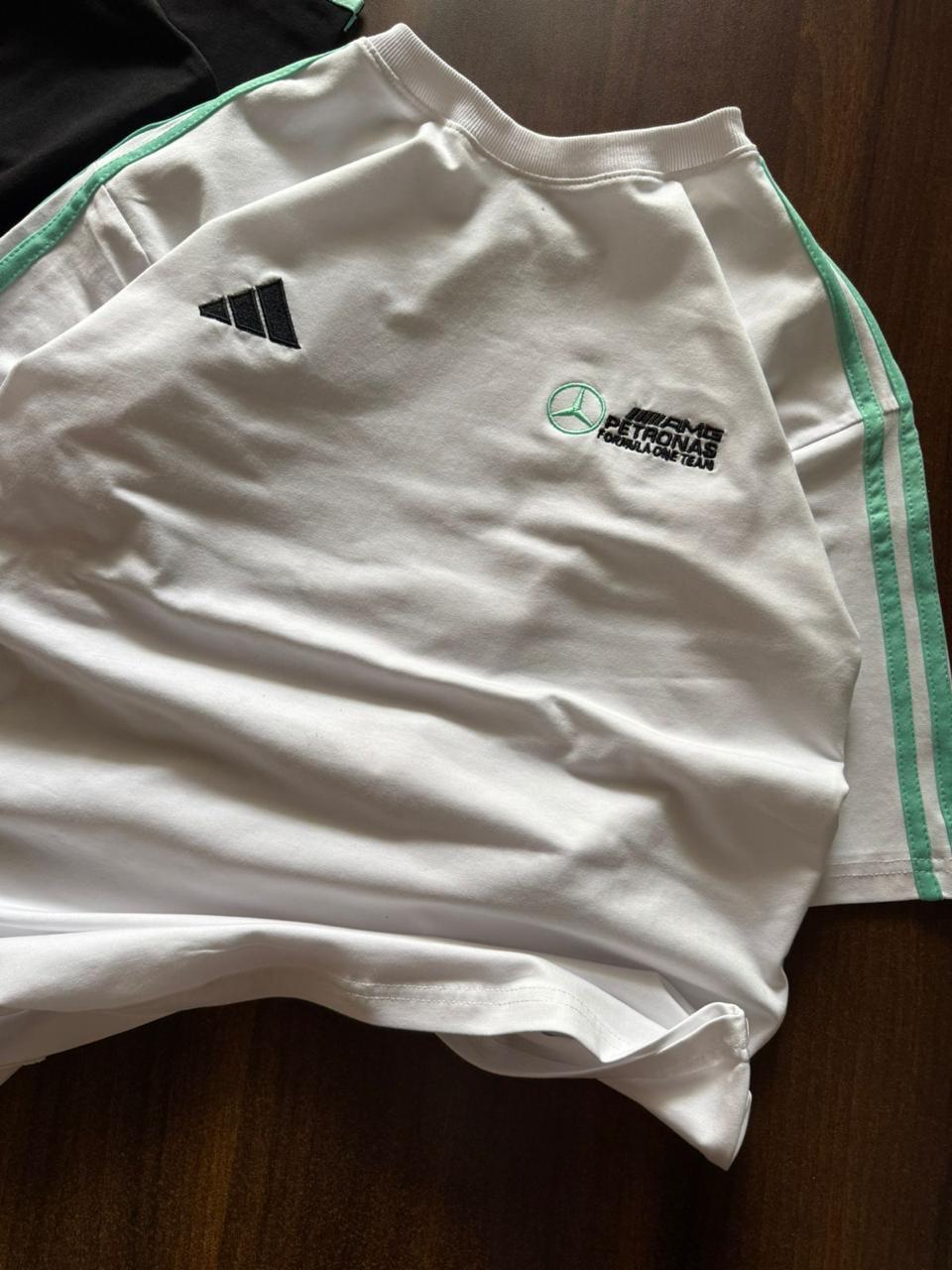 A white Adidas Mercedes-Benz Campus Team t-shirt laid flat on a dark wooden surface, showing the black three-stripe logo and the green and black Campus Team chest graphic.