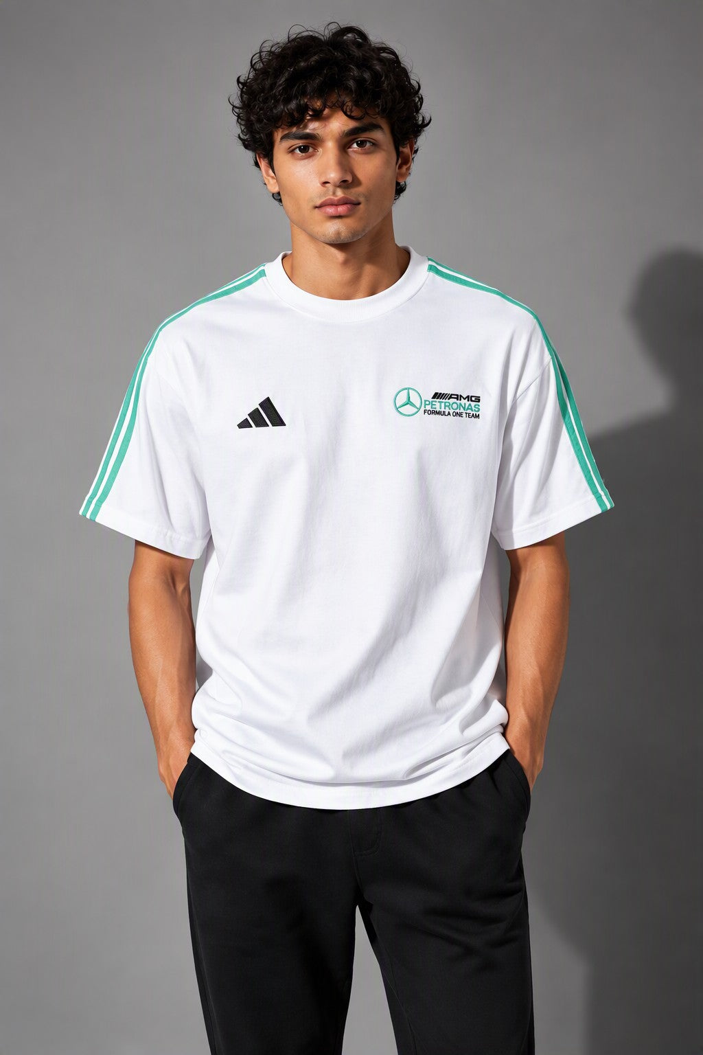 A man wearing a white Mercedes-AMG F1 team Adidas t-shirt with green shoulder stripes and logos, paired with black pants, standing against a gray studio background.